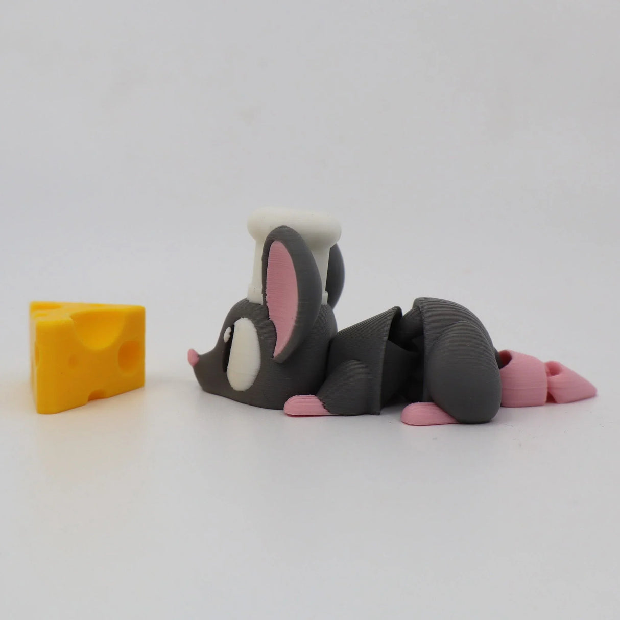 3D printed gray mouse toy with chef hat lying by yellow cheese wedge, sensory toy