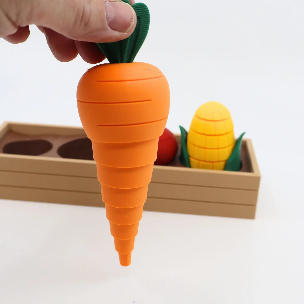 Hand holding a 3D printed sensory carrot toy, with corn and wooden box in background.