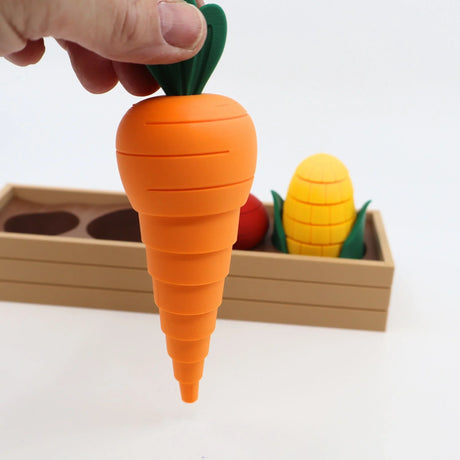 Hand holding a 3D printed sensory carrot toy, with corn and wooden box in background.